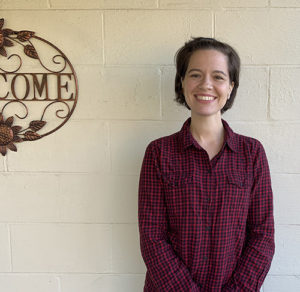 Montessori teacher, Milena Thomas, standing outside her school building smiling, wearing a plaid button down shirt. A sign next to her reads "Welcome".